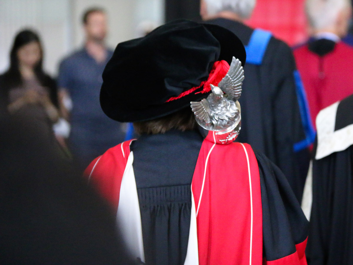 The Clerk of Senate walks towards the stage in full regalia while holding a silver mace over her shoulder.