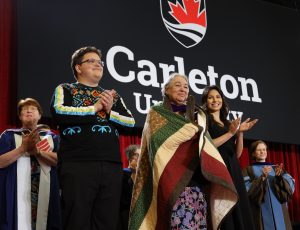 Three people stand on stage at Carleton's Convocation clapping