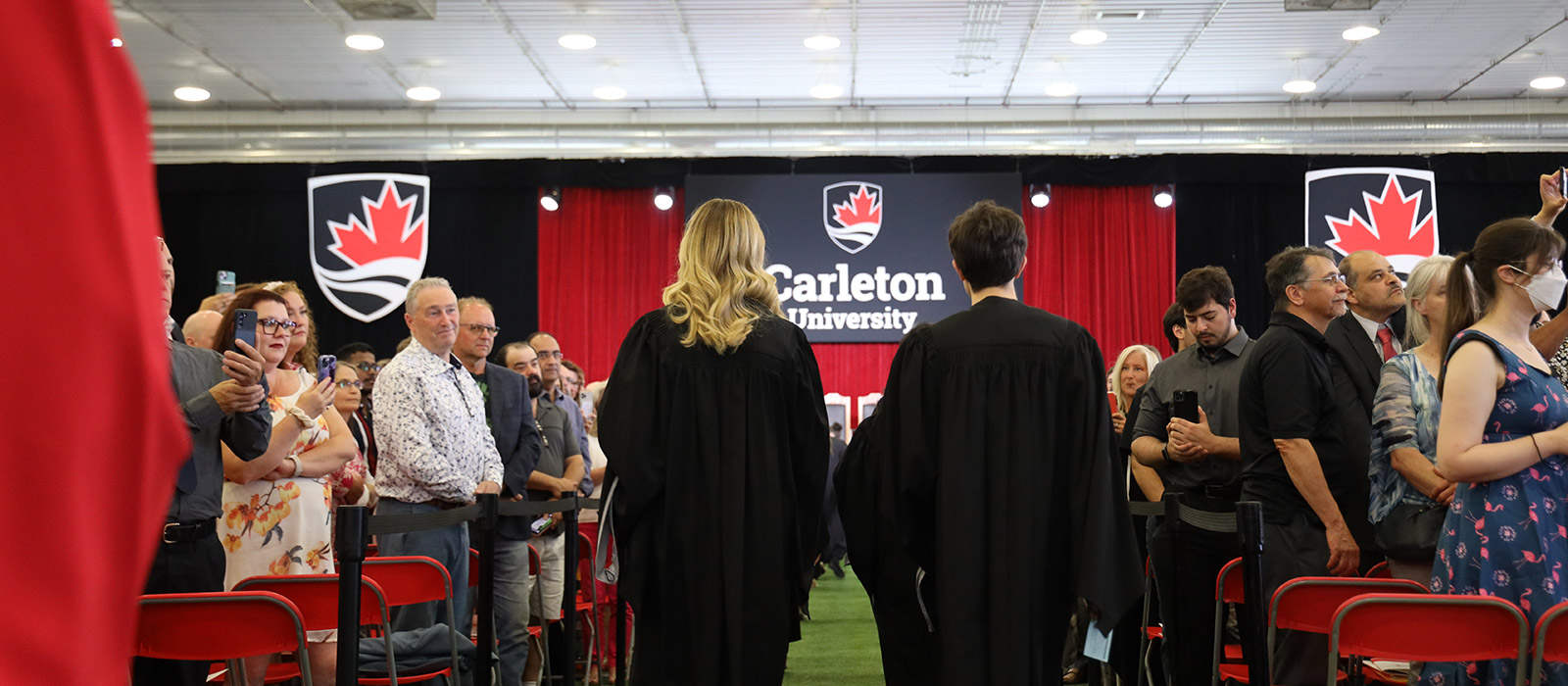 Two graduates makes their way towards the stage down a row with crowds on both sides.