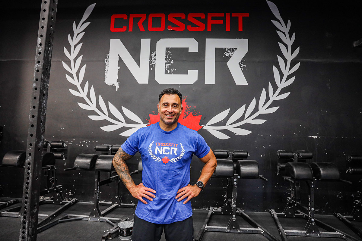 A man smiles for the camera with his hands on his hips, while standing inside a CrossFit gym.