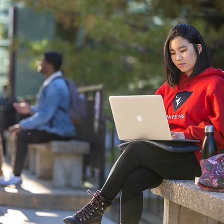 A Carleton University student wearing a red Ravens hoodie sits on a bench using a laptop