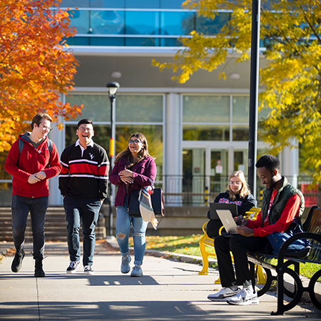 Three Carleton University students walk together on campus while two other students sit on benches using laptops.