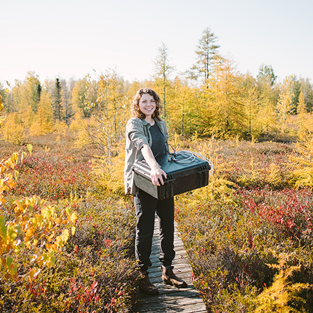 A young woman holds a piece of machinery whole standing on a pathway in a peat bog