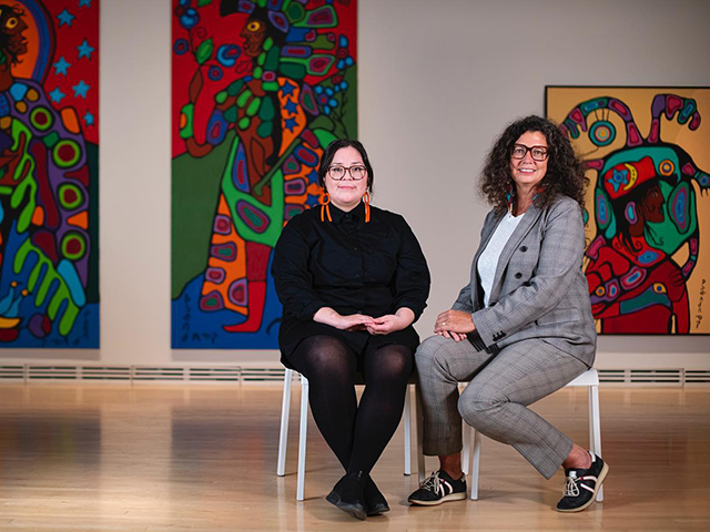 Two people pose for a picture while seated in front of an art exhibit.