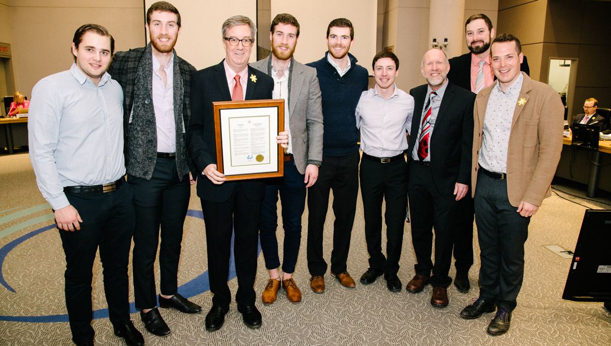 The men's curling team gathers with Mayor Jim Watson and Councillor Mathieu Fleury in council chambers while holding a framed declaration.