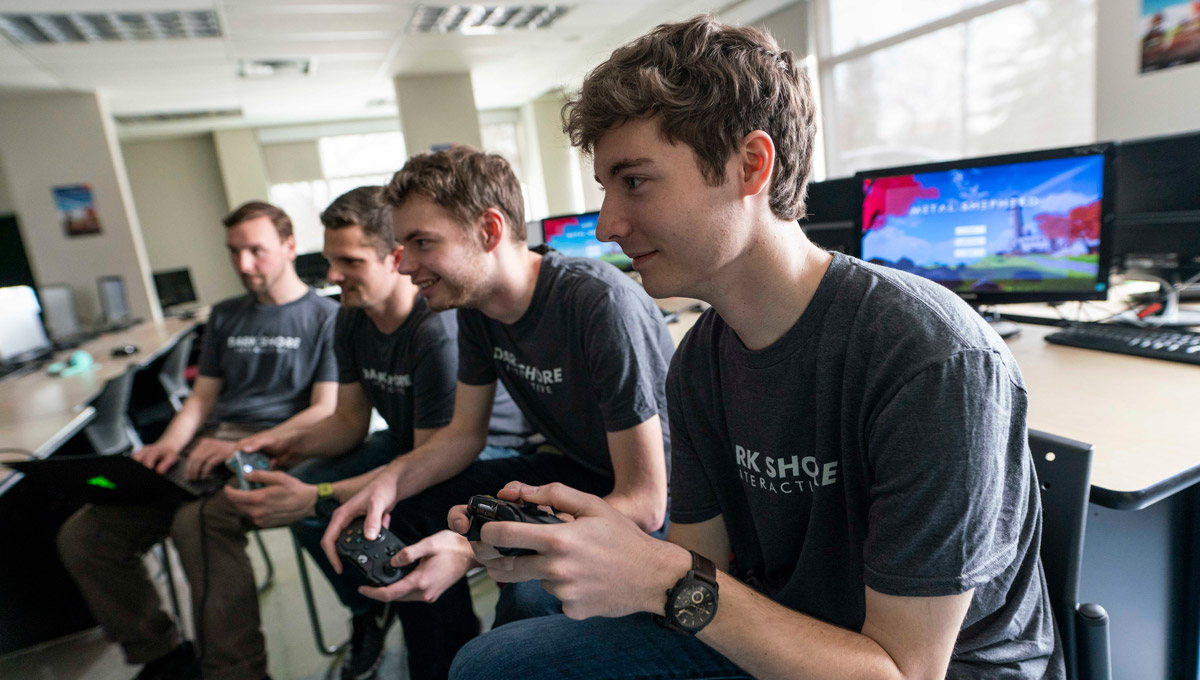 A group of students wearing shirts with the name Dark Shore Interactive on them play a video game using controllers.