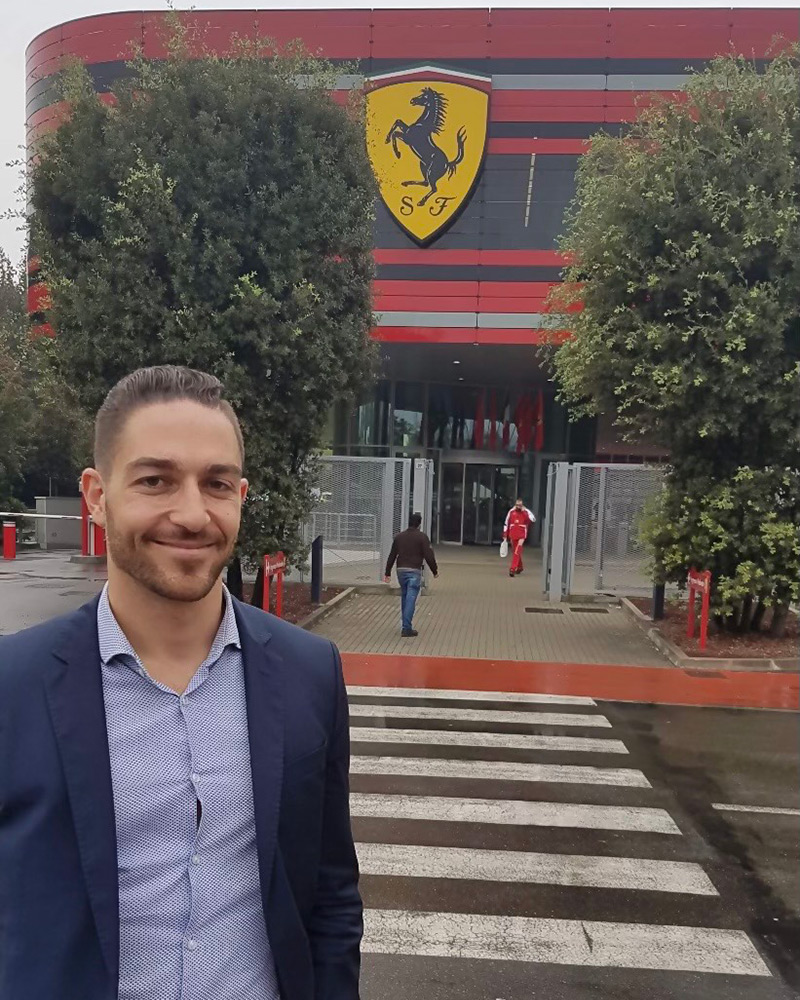 A man in a dress suit poses for a photo with the Ferrari logo visible in the background.