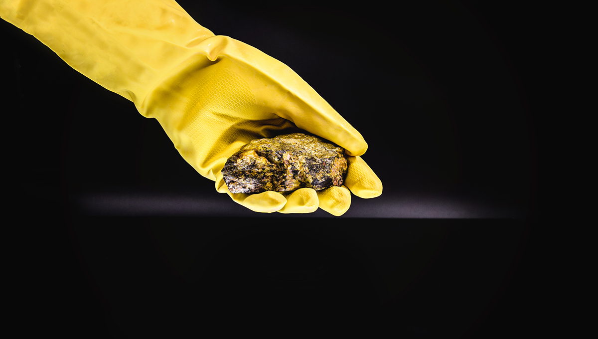 A hand in a yellow protective glove holds a yellow and black-looking rock