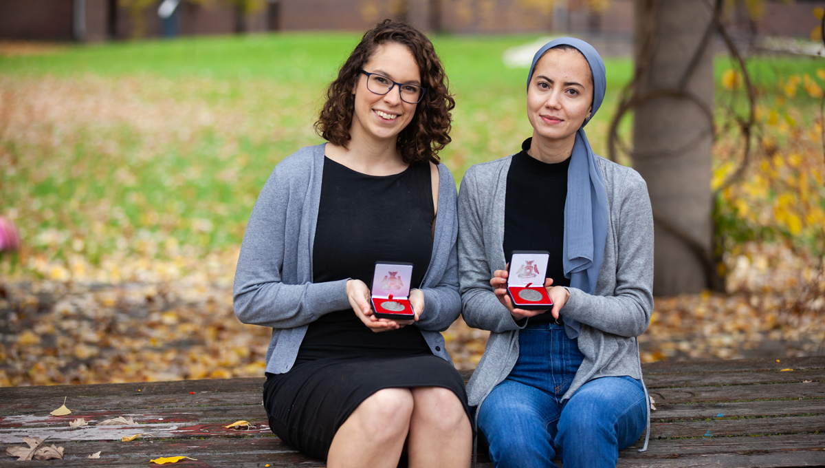 Mercedes Veselka and Samah Saci hold their medals outdoors on a bench in Alumni Park with fallen leaves in the background.