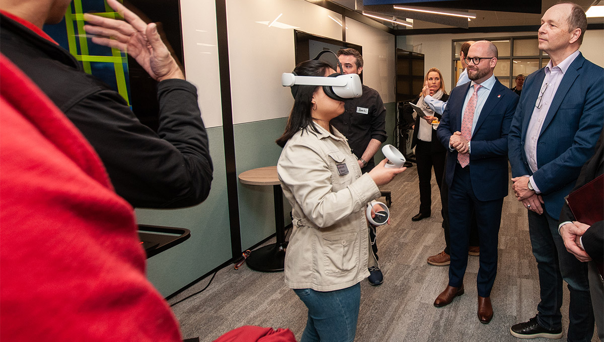 A small crowd watches a woman demonstrate virtual reality while wearing a VR headset and holding VR controllers.