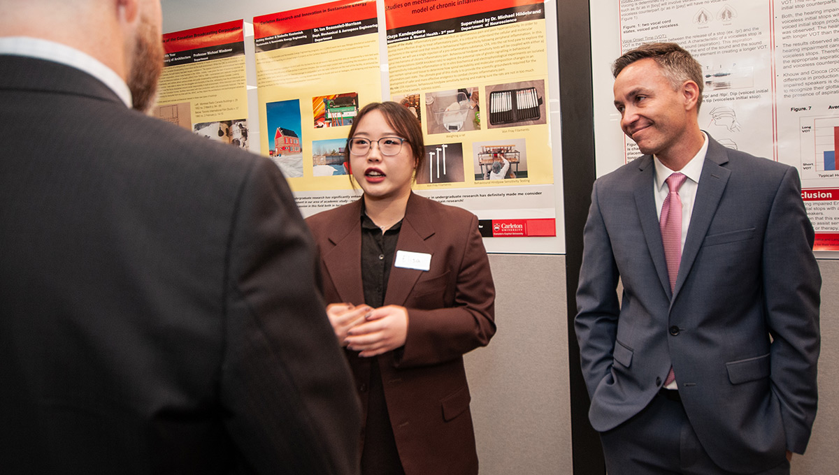 A woman wearing glasses and a brown blazer engages with people about the work featured behind her.