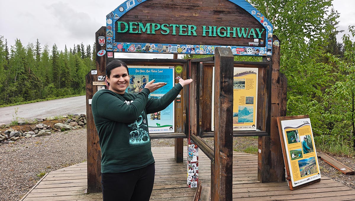 A woman wearing a dark green sweater points at a sign that says Dempster Highway, on the way to supporting remote communities.