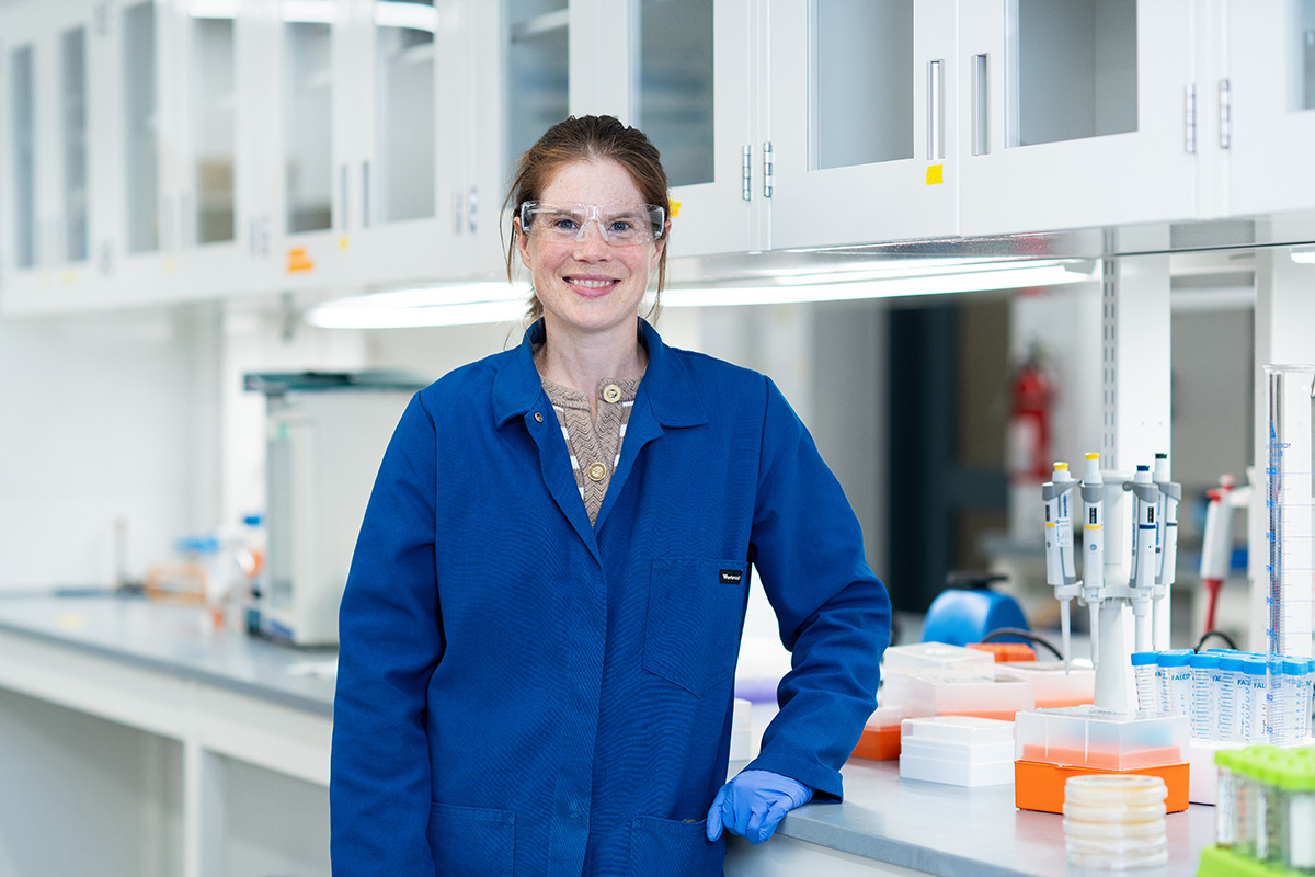 A woman wearing a blue lab coat and safety goggles smiles for the camera while leaning against a counter.