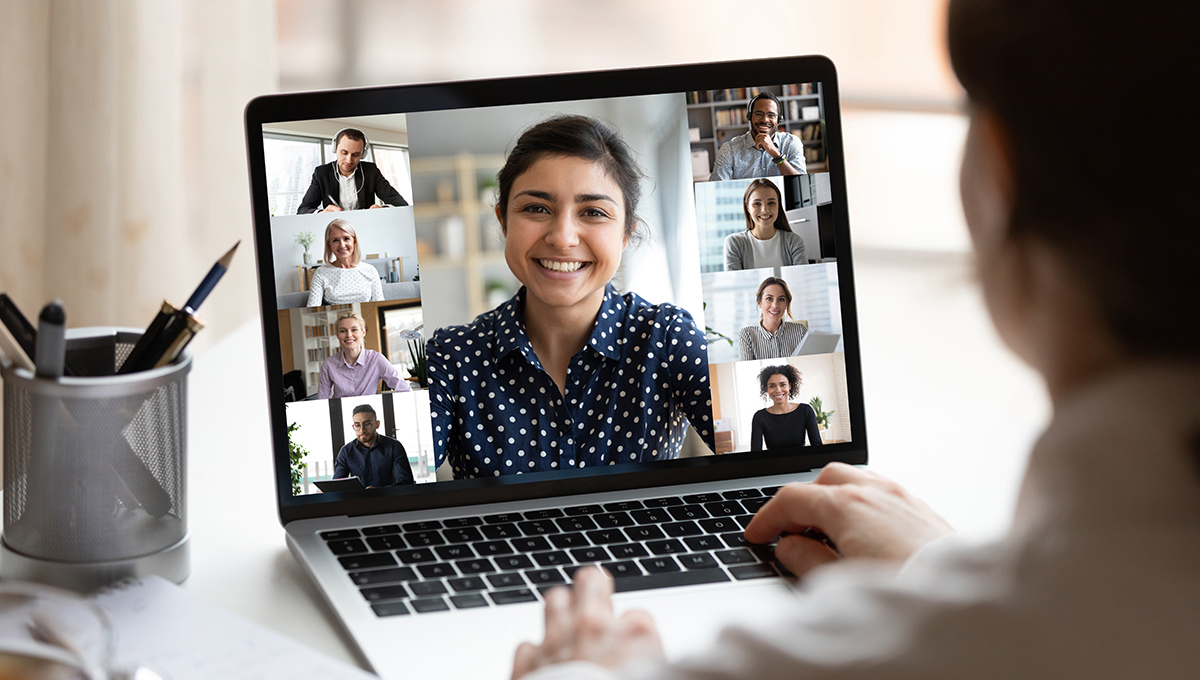 A woman sits at a desk looking at computer screen where a collage of diverse people can be seen in video conference