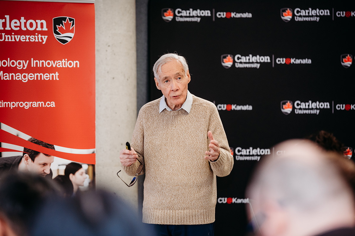 An older man with a beige sweater, holds his glasses in his hand while addressing an audience.