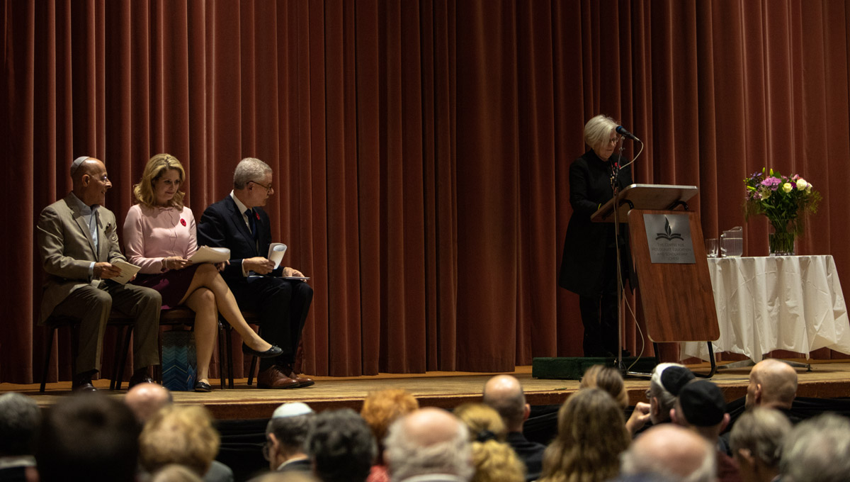 CHES Director Mina Cohen stands at a podium and speaks into a microphone while a group of three panel members look on.