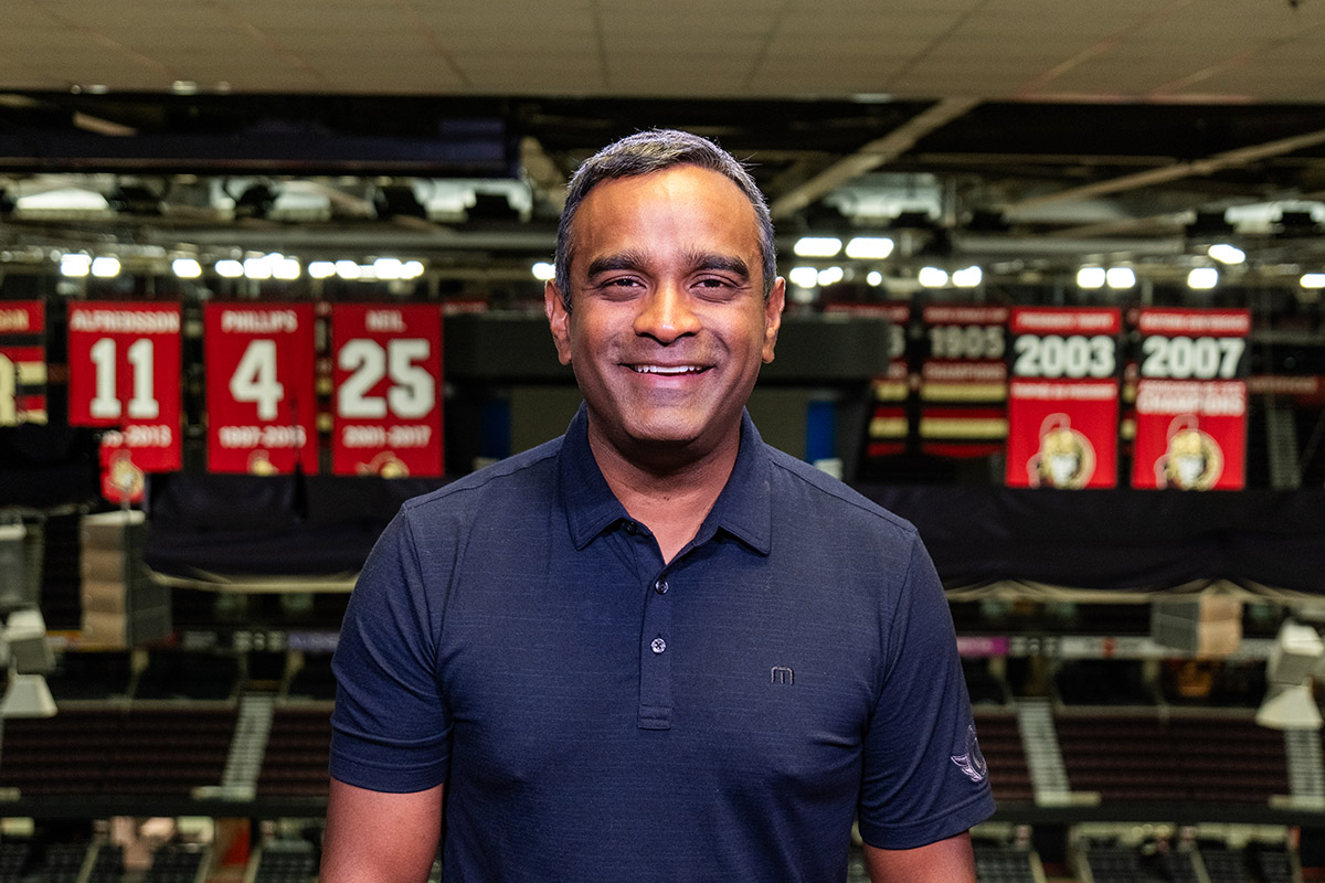 A man wearing a navy blue golf shirt smiles for the camera with banners hanging from the arena rafters behind him.