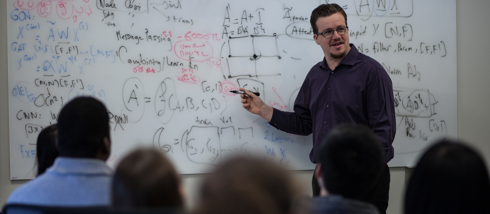 A man with glasses stands points at a white board with formulas on it.