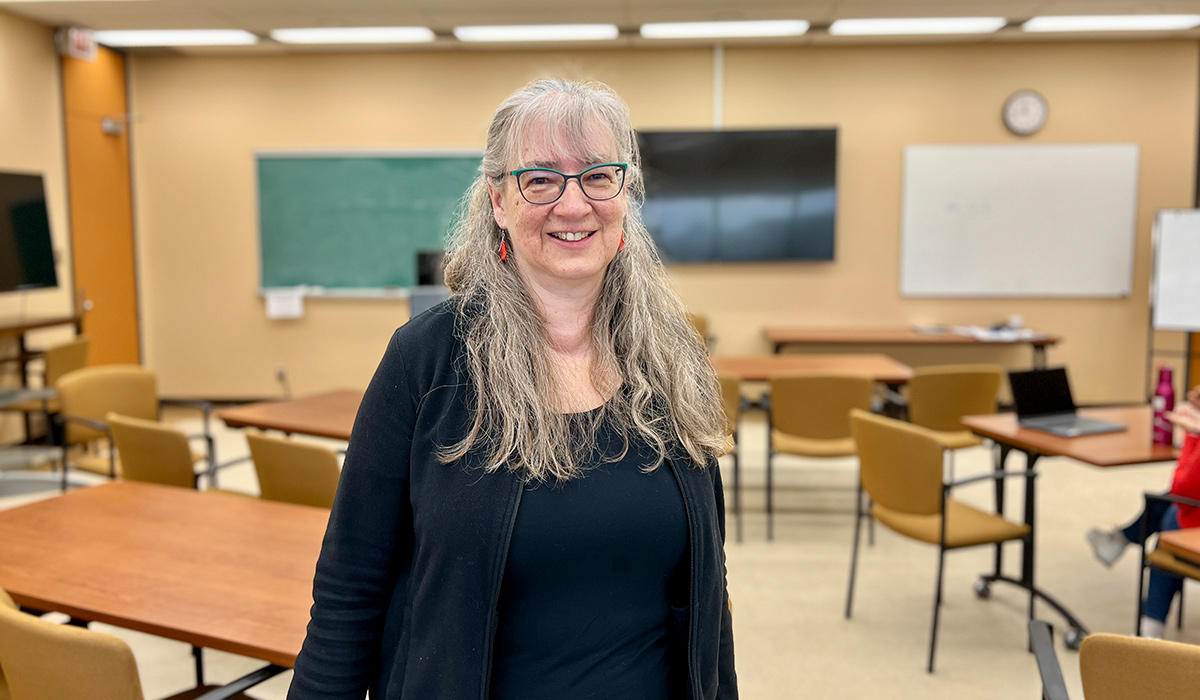 A woman wearing glasses with long, light hair smiles for the camera while standing in a classroom.