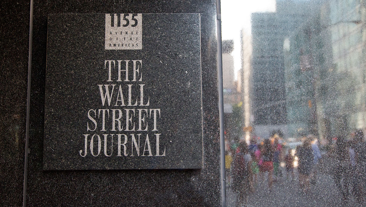 Photo of the sign of The Wall Street Journal outside of its building. At right there are some people and buildings reflected by the marble.