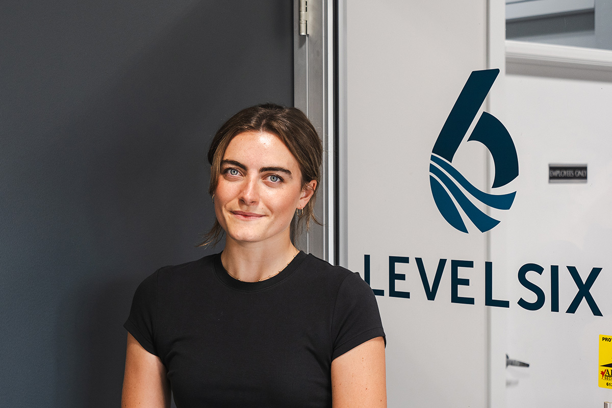 A young woman wearing a black shirt poses for a photo next to a wall with the Level Six logo