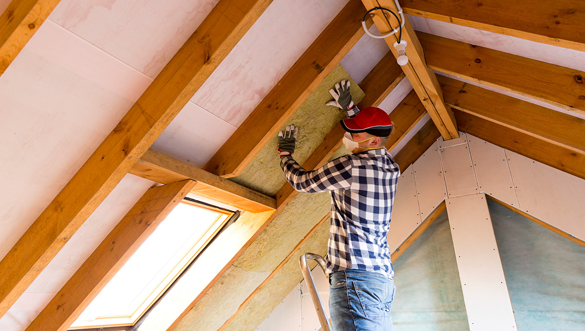 A construction worker stands on a ladder to install insulation on the interior of a roof.