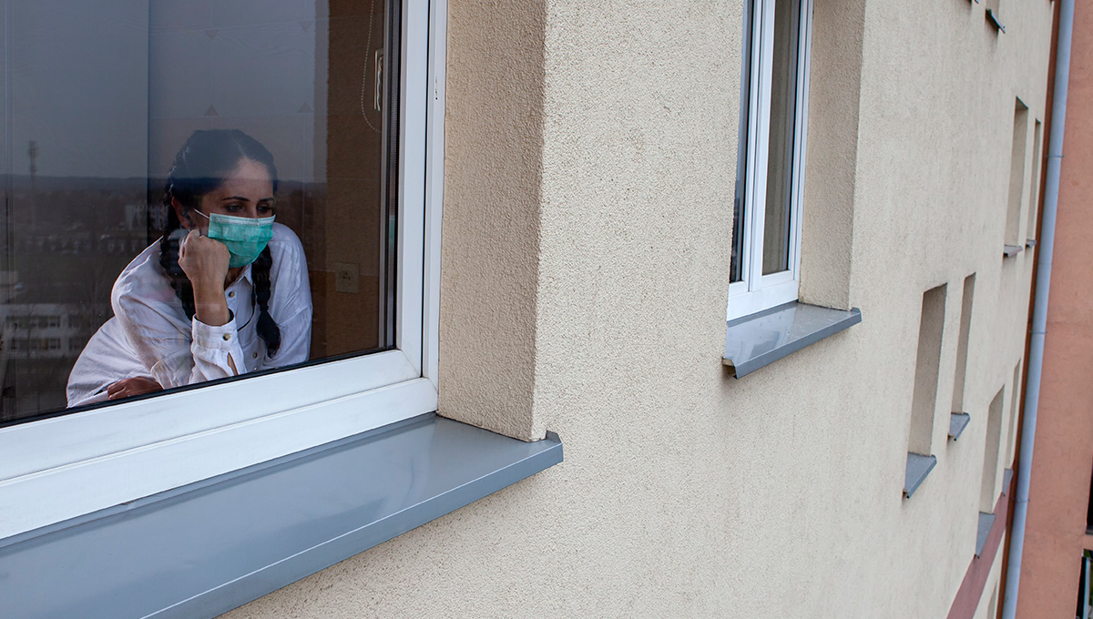 A 40-year-old woman wearing a protective mask is looking out of the window. Home quarantine for 14 days due to the coronavirus COVID-19 epidemic.