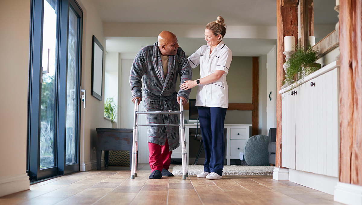 Senior Man In Dressing Gown Using Walking Frame Being Helped By Care Worker