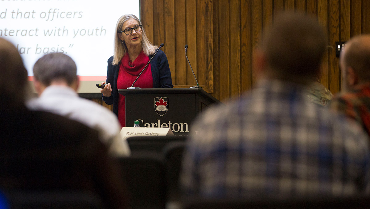 A blonde woman with glasses and a red shirt speaks from a podium