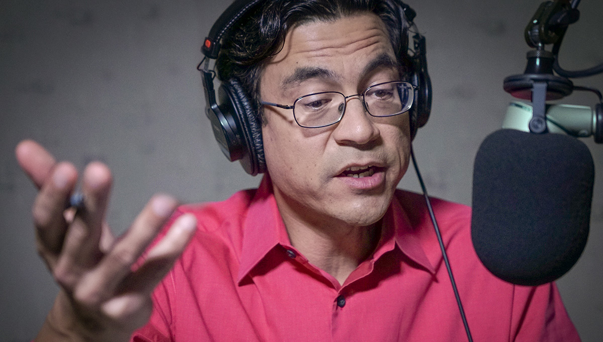 A man in glasses wearing headphones speaks passionately into a microphone.