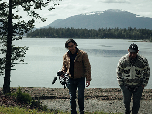Two men, including one holding a camera, walk away from the shore of a lake.