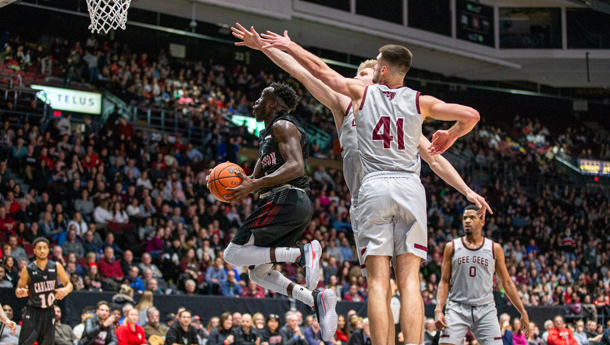 A Ravens men's basketball player goes to the basket against two uOttawa Gee Gees players.