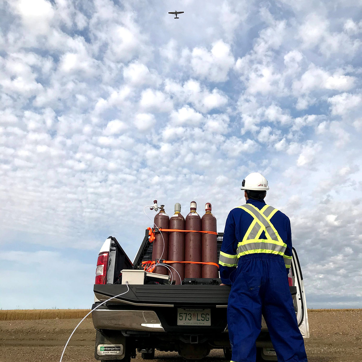 A man standing behind a truck looks up at an airplane overhead