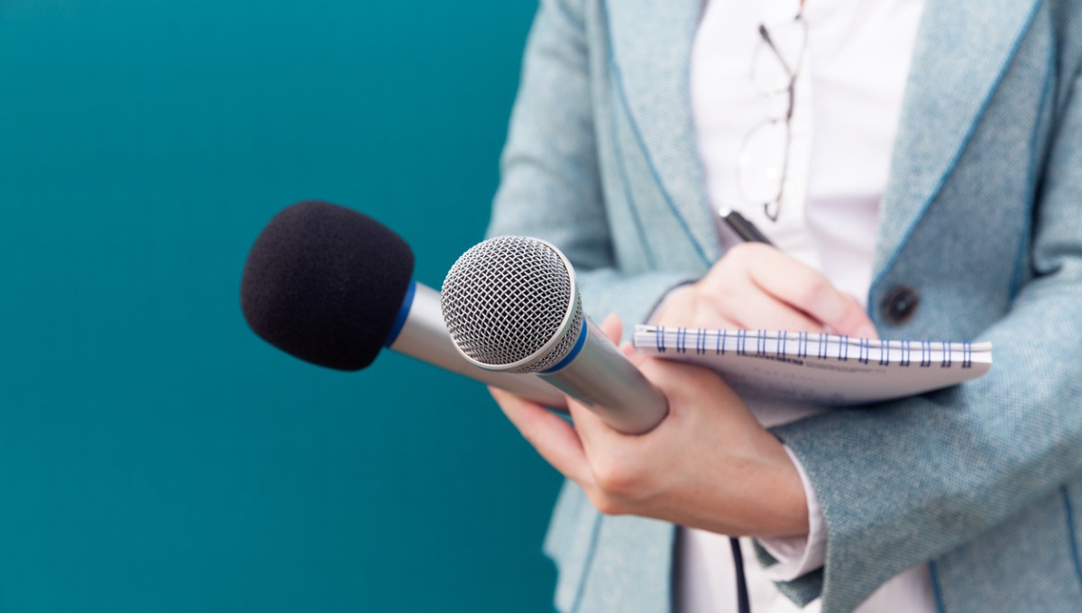 Close up of female news reporter's hands holding microphones while also writing in book