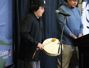 A young man in a black shirt plays a hand drum on stage in front of a microphone as another man listens with his eyes closed.