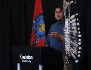A man with a blue hoodie speaks into a microphone behind a podium with the words 'Carleton University' written on the front.