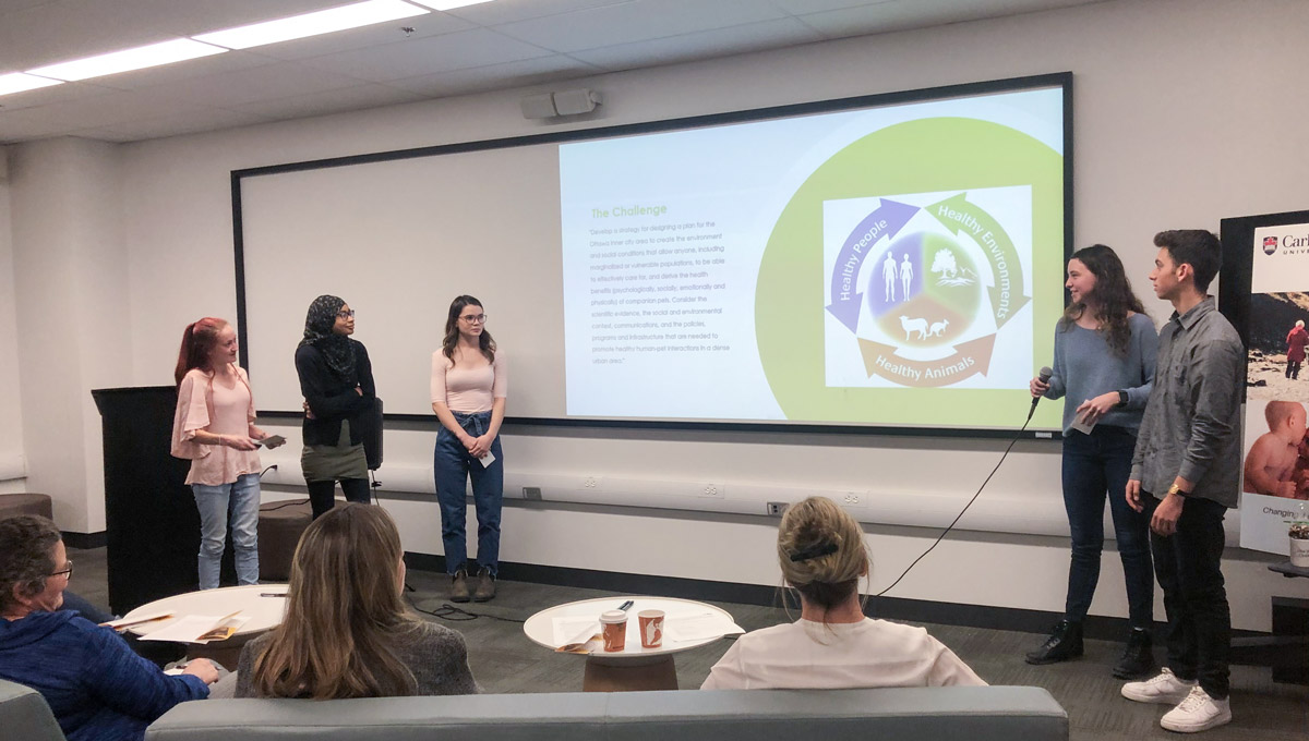 A students holds a microphone while delivering a presentation during the One Health Challenge, with her collegues looking on..