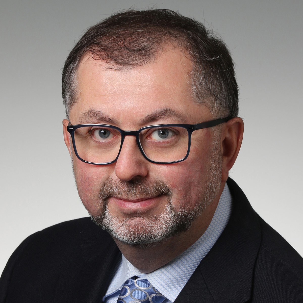 A professional headshot of a man wearing glasses and a dark suit.