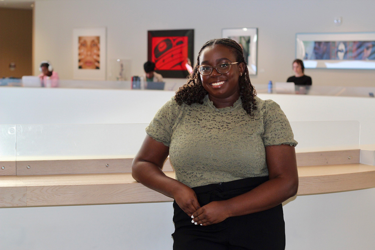 A young woman with glasses leans against a railing while smiling for the camera.