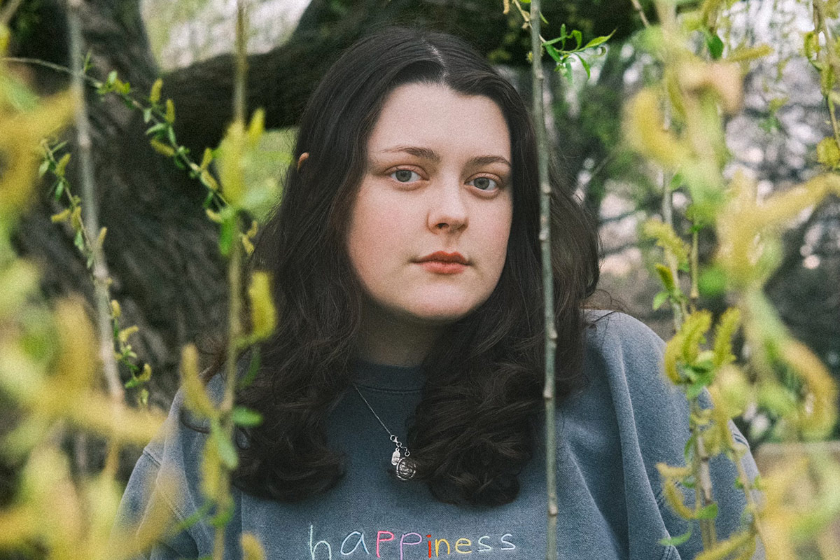 A photo of a young woman with long brown hair, sitting among plants.