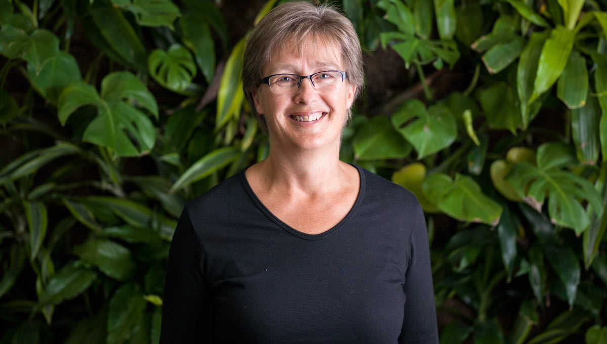 A woman with short hair and glasses in a black long-sleeved shirt stands in front of a wall of plants
