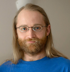 A portrait of a man in a blue shirt with glasses and long blonde hair.
