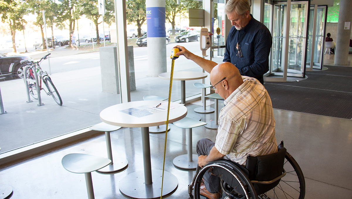 A person in a wheelchair and another man measure a table to meet Accessibility Certification rules.