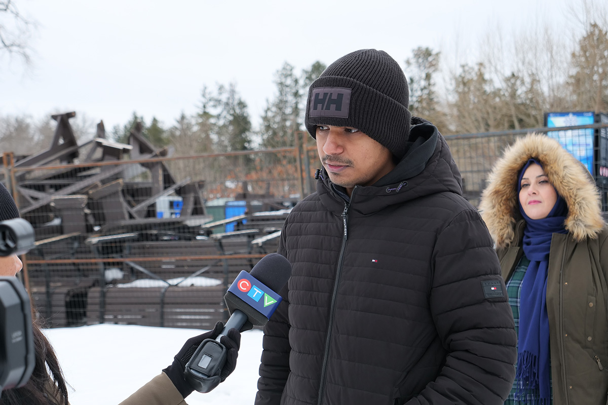 A man in a black jacket and a black toque speaks into a journalist's microphone.