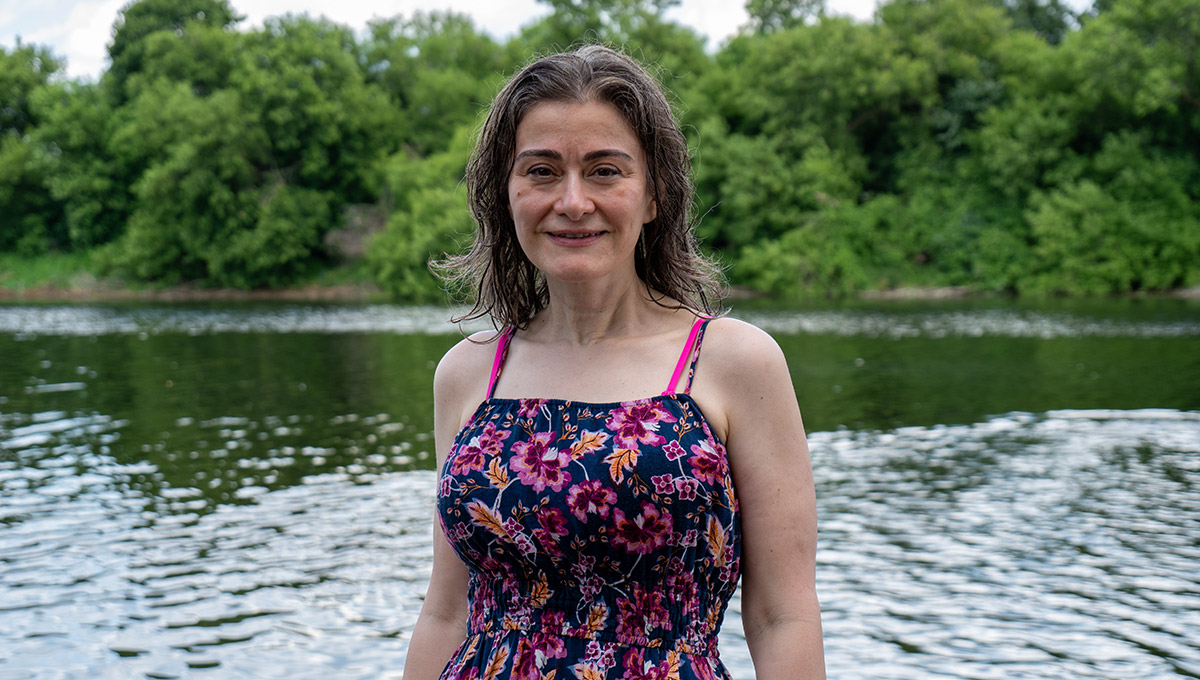 A woman wearing a floral bathing suit poses for the camera in front of a river and trees while demonstrating the water quality of the Rideau River.