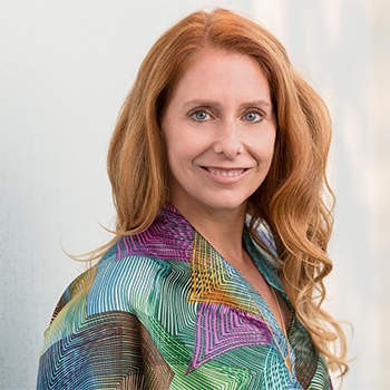 A professional headshot of a woman with red hair wearing a multi-coloured shirt.