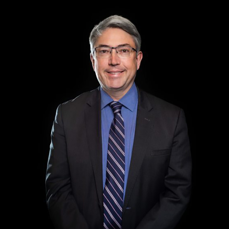 A professional headshot of a man with glasses wearing a dark suit and blue dress shirt.