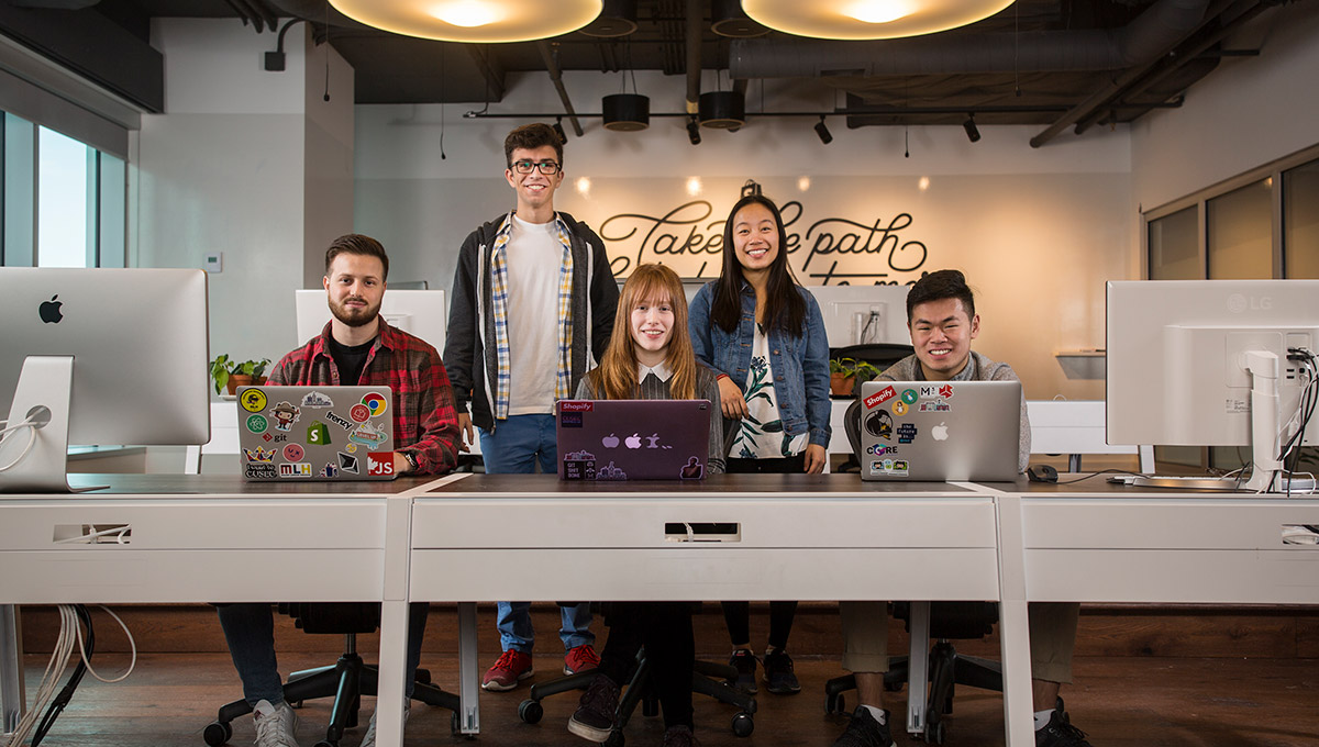 Students in Carleton's Dev Degree partnership with Shopify (pictured here at computers in the Shopify headquarters) split their time between campus & the company’s headquarters, earning valuable career experience.