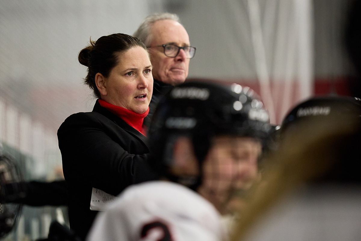 A hockey coach stands behind her players on the bench during a game.