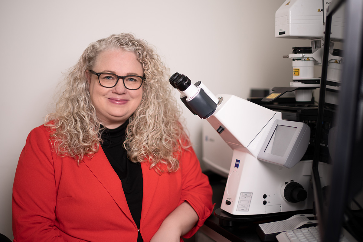 A woman with long blonde curly hair, and glasses, wearing a red dress jacket, smiles for the camera while sitting next to a microscope.